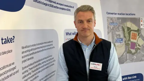 Connor Bennett/BBC A man with grey hair and brown eyes is wearing a blue shirt and navy gilet with a brown collar. Pinned to the gilet is a name tag which reads LionLink Gareth construction director. He is standing in front of an information display showing blocks of text and a map.