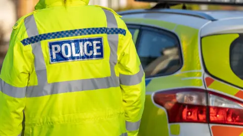 The back of a police officer in a fluorescent yellow jacket, with the word POLICE printed on the jacket. In front of the officer, is a marked police car, which is yellow, blue and orange. 
