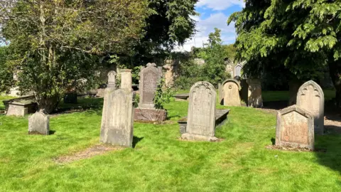 Old gravestones, some of the leaning back or to the side, in a grassy area surrounded by trees at Chapel Yard cemetery.