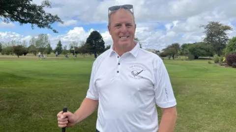 Andy Wilkins - a man with grey hair and sunglasses on his head - smiling. He is wearing a white polo shirt with an emblem of a golf club. He is holding a golf club. He's standing on the golf green with golfers behind him and a flag and some trees