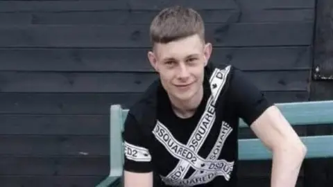 A young man with short brown hair wearing a back t shirt with white bands across it and the text Squared2. He is sat on a blue wooden bench with a shed behind him and is smiling at the camera.