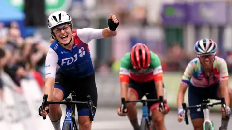 Grace Brown of team FDJ Nouvelle-Aquitaine Futuroscope celebrates as she crosses the line to win stage four of The Women's Tour from Wrexham to Welshpool on 2022. She is wearing a blue and white cycling suit, glasses and a white helmet. Her arms is raised in a fist as she rides the bike.