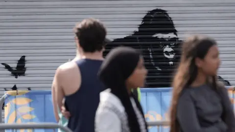 PA Media A man looks at Banksy's artwork of a black gorilla on a silver metal shutter, as women walk past unfocused