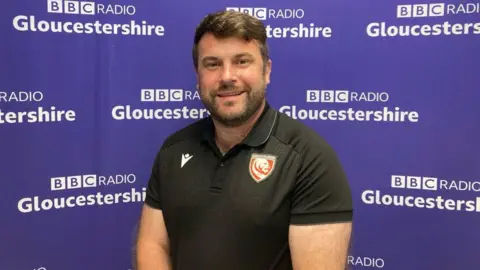 A man with brown hair and a beard wearing a black Gloucester Rugby top. He's stood in front of a purple board which has 'BBC Radio Gloucestershire' printed across it.