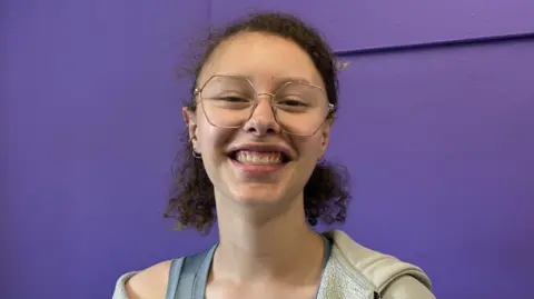 A young woman has brown curly hair tied back in a short ponytail, she is wearing large gold frame glasses and is smiling. She is wearing a grey vest and a grey hoodie. She is sitting in front of a bright purple wall