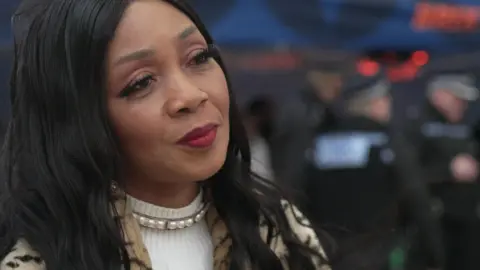 Pastor Lorraine Jones Burrell is looking to the right. She has long black hair, wearing a pearl necklace over a white turtle neck top and a animal print jacket. The background is blurred, but shows police officers in black uniforms stood away from her, with their backs to her.