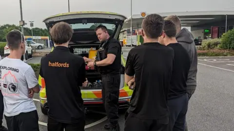West Mercia Police A police officer dressed in black standing in front of his police car, which is parked in a petrol garage with the boot open. He is talking to a group of young men who have their backs to the camera.