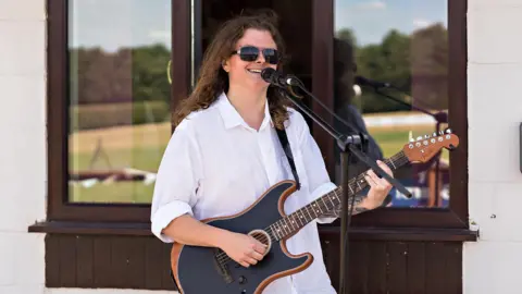 Ben Smith Music Ben Smith, who is wearing a white shirt and dark sunglasses, playing a guitar and singing into a microphone at an outdoor event.