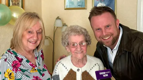 Mrs Taylor stands centre smiling at the camera, wearing a patterned cream, brown and grey woollen cardigan and glasses. A woman stands to her right wearing a flowery and striped shirt. Brett stands to her left with his arm around her shoulder and a purple microphone with BBC Radio WM written on it in his left hand. There are green and yellow balloons, a floor lamp and a few pictures on the wall behind them.