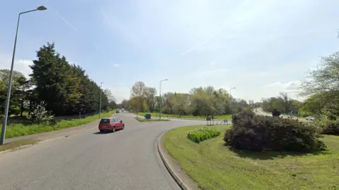 A roundabout with grass to the right. A red car is going around it. Ahead is a long road lined with trees. It is a sunny day.