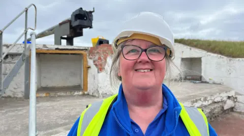 Diane Stephens, a blonde-haired woman stands in front of the camera smiling, wearing a white hard hat, blue fleece and fluorescent tabard.
Behind her is a World War One gun battery.