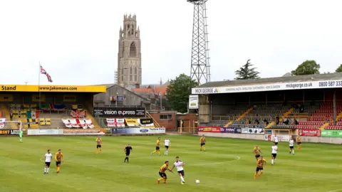 A football match is being played in a small stadium with yellow, black and seating. One team is wearing a yellow and black striped kit, the other is wearing white. The Boston Stump, a church tower, is visible behind the stands. Floodlights are visible in the background and a flags and banners are also hanging in the stands behind the goal.