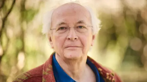 Philip Pullman with glasses in a brown & beige cardigan and blue shirt against trees and leaves background
