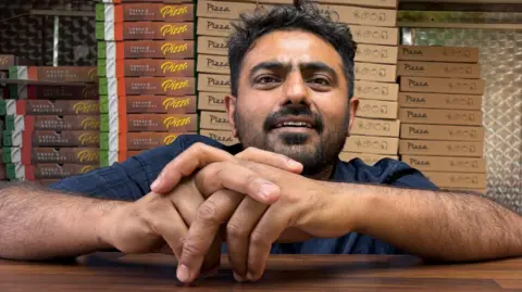 Martin Heath/BBC Iyaz Malik with short dark hair leaning on a wooden takeaway counter inside a pizza shop, with stacks of pizza boxes piled high behind them and a metallic kitchen wall with red emergency switches in the background.