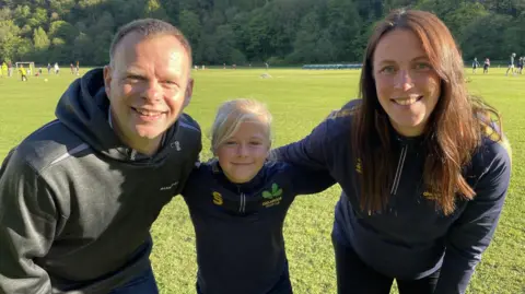 Edie, nine, stands smiling between her dad Matt and her coach Amy Sherratt on the grass at Oakamoor Cricket Club. She wears a navy Oakamoor Cricket Club training top with the club's oak leaf badge. Behind them, junior players take part in a training session against a backdrop of wooded hillside on a bright, sunny evening.