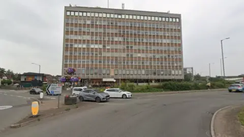 Google A nine-storey building, which is brown with windows, is in the background. Part of a roundabout is visible on the far right and cars are about to enter the roundabout from a road on the left. 