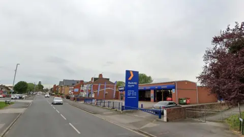 Google A Google Street View of Hull Road, Anlaby. There are paths on both sides of the road. A Kwik Fit branch is on the right and there are houses in the distance.