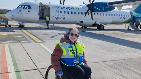 Southampton Airport Hope wears a high vis vest sunglasses. She is sitting in a wheelchair with an air lingus plane behindat Southampton Airport. 