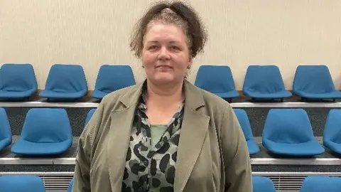 LDRS Amanda Napper had long brown hair that is tied up. She is wearing a khaki coloured jacket and black and white spotted top. She is smiling.