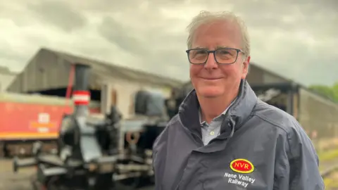 Chairman standing in front of a locomotive while wearing a grey rain coat with the red and yellow branding for the railway. Mr Purcell is smiling at the camera, has square framed glasses and short white hair. 