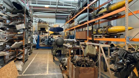 Historical RAF items including bombs, parts of planes and various other equipment in a warehouse. Some are on shelves while others are in boxes.