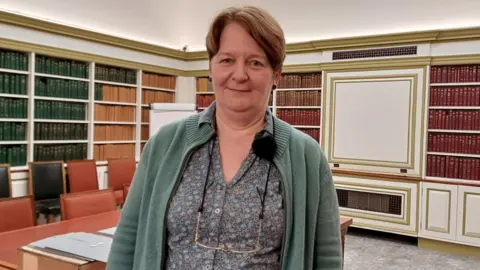 Ulrike Hogg is wearing a floral pattern blouse and a green cardigan. She is smiling as she stands in the national library. There are shelves filled with books behind her.