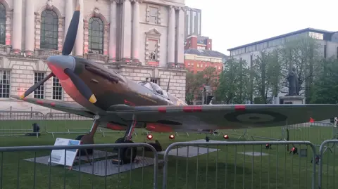 A replica Supermarine Spitfire, used extensively by the Allied Forces during World War Two, on display on the grass outside Belfast City Hall, which is partially visible in the background. 