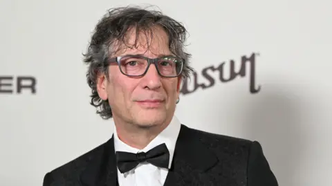 Neil Gaiman at a gala in Los Angeles, California, US. He is wearing a suit and bow tie and is looking at the camera 