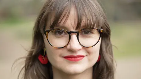 A woman wearing glasses smiles at the camera. She has dark hair, a fringe and wears red lipstick