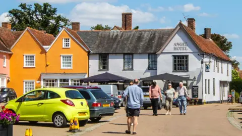 Getty Images An image of the outside of the Angel Hotel, in Lavenham from the road. It is next to a bright orange building. People can be seen walking to and from the hotel. There are cars parked up to the left of the image 