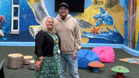 Robbie Meakin A man wearing a hat, beige hooded top and jeans stands next to a woman wearing a black cardigan and long green dress. Behind him is a children's play area, with colourful murals of the sea and animals on the walls.