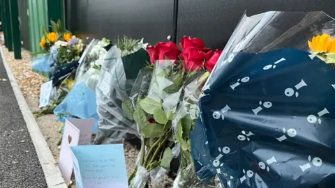 George Carden/BBC Algunas alcantarillas y tarjetas de flores se fueron para el difunto Billy Vigar en el Chichester City Football Club. Se pueden ver seis alcantarillas de flores separadas continuamente con tres cartas. 