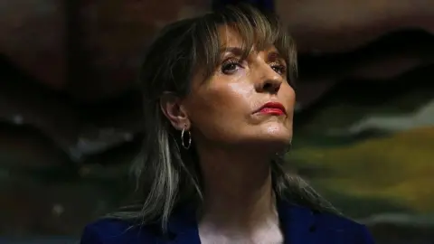 A woman with shoulder length brown hair is wearing a blue jacket 