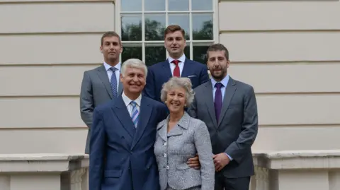 Contributed The Young family stand together in front of a white building. The three children, who are men, stand behind their parents. They are all wearing smart and formal attire. They are smiling at the camera.
