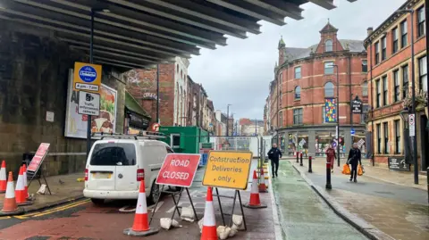 The entrance to a road under a bridge which has three signs in place, one which reads 'Road closed'. There are several orange and white cones placed around the signs and a parked white van. In the background are shops and people. 