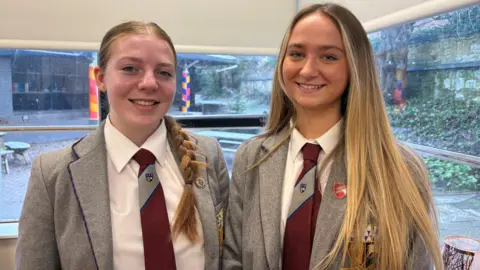 Connie and Sarah smile in their gray school uniforms. They both wear badges and wear maroon and brown ties with white shirts.