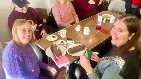 A group are sitting round a table with two birthday cakes, two of the women at the front are smiling at the camera