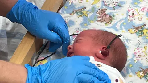 a very small baby with an earbud in its ear, connected to a long cable going over his head. Gloved hands are holding the cable and the baby