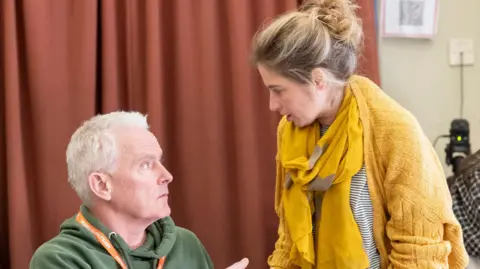 Johan Persson A man with short grey hair and a green jumper speaks to a woman with tied up blonde hair wearing a yellow cardigan and scarf.