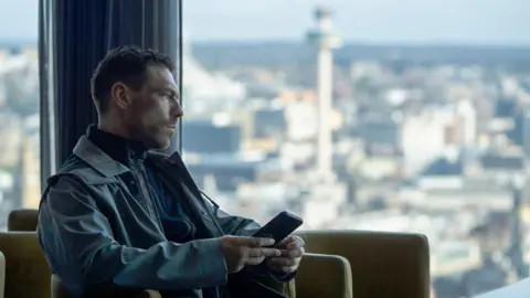 BBC/Left Bank Pictures/James Stack Gang member Michael Kavanagh (James Nelson-Joyce) with brown hair wearing a blue coat and navy sweater zipped up in a scene from This City Is Ours. He sits in a high rise building holding a phone with large windows showing an aerial view of Liverpool city centre.