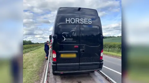 Alexandra Jones A black horsebox with the word 'Horses' on it, written in white. Below is a logo of a horse and the words 'Southwest Horse Boxes'. The vehicle is on a flatbed truck, about to be towed away and there is a man stood to the side. The truck is parked on the side of the road, with green fields either side. 