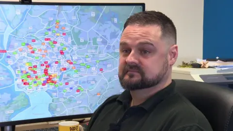 John McVicar sits in front of a large digital map which shows taxis around Plymouth. He is wearing a black polo shirt.