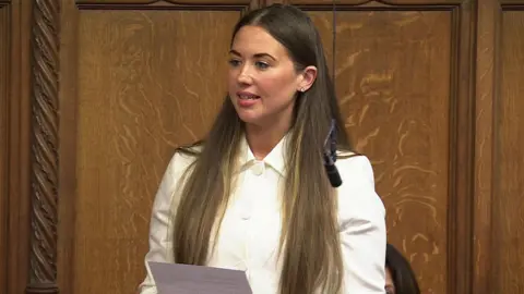 HOC Sorcha Eastwood - a woman with long brown hair wearing a white blazer delivers a speech in the House of Commons