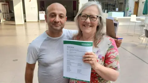 Codford Tea Rooms Kevin and Lizzie smile at the camera from the Wiltshire Council foyer area. They are holding Wiltshire Council branded paper to the camera showing the meeting agenda