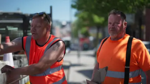 Two men wear high-vis orange clothes as they work by the side of the road