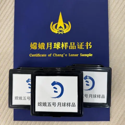 Mahesh Anand Uma pasta azul que contém uma certificação da agência espacial chinesa sobre as amostras Chang'e 5. No topo estão três caixas com fundo branco e bordas pretas. Na frente está o logotipo da agência espacial chinesa Chang'e e a redação do roteiro chinês.