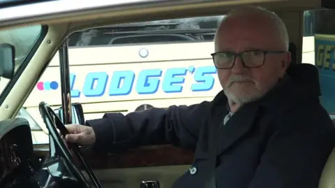 Bill Adams sits in the driver's seat of his Rolls Royce. He has one hand on the steering wheel and is facing the camera to his left and smiling. He wears a navy coat and glasses. He has short grey hair and a grey beard.