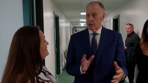 Sir Geoff Hurst gesturing with his hands and talking to the head teacher during a visit to the school. He is wearing a suit and standing in a corridor.