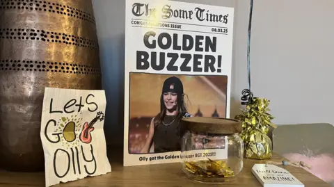 A side table in a living room with a bronze coloured vase, a mock newspaper front page with a picture of Olly and the headline Golden Buzzer and another card that reads Let's Go Olly.  There is also a clear glass jar that contains gold confetti from the Britain's Got Talent Show. 