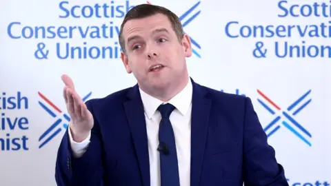 A man with dark, wearing a dark blue suit and tie, speaks with his right hand raised in front of him. He is standing in front of a Scottish Conservative-branded background  